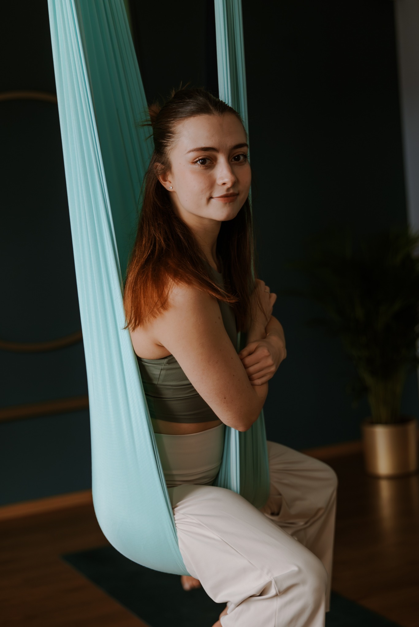 Kobieta w odzieży sportowej siedzi w hamaku do aerial jogi, uśmiechając się do kamery. W tle ciemna ściana i roślina w złotej doniczce.