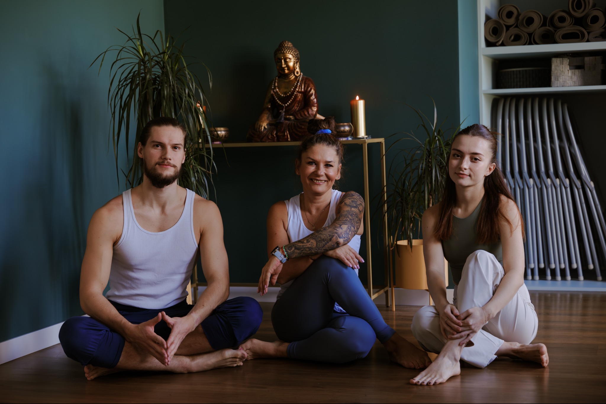 Troje uśmiechniętych ludzi siedzi po turecku na podłodze w studio jogi z posągiem Buddy i matami w tle. Kobieta z tatuażem na ramieniu w centrum.