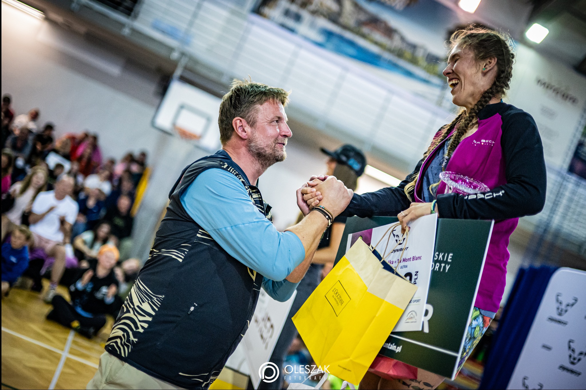 Trener biegania gratuluje zawodniczce sukcesu po zawodach. Uśmiechnięta kobieta trzyma nagrody i upominki. W tle widownia i sala sportowa.