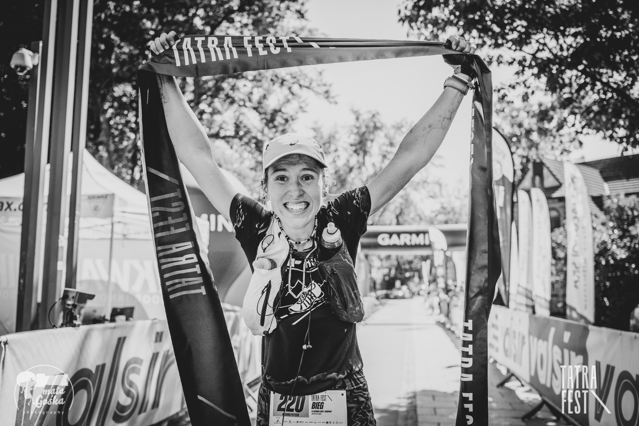Uśmiechnięta kobieta z numerem 220 na mecie biegu Tatra Fest, z podniesionymi rękami trzyma baner. Czarno-białe zdjęcie z widocznym logiem Garmin w tle.