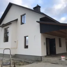 Dom jednorodzinny z białą elewacją i ciemnymi elementami wykończeniowymi, widoczna drabina i wiadro farby, sugerujące świeżo wykonane prace elewacyjne.