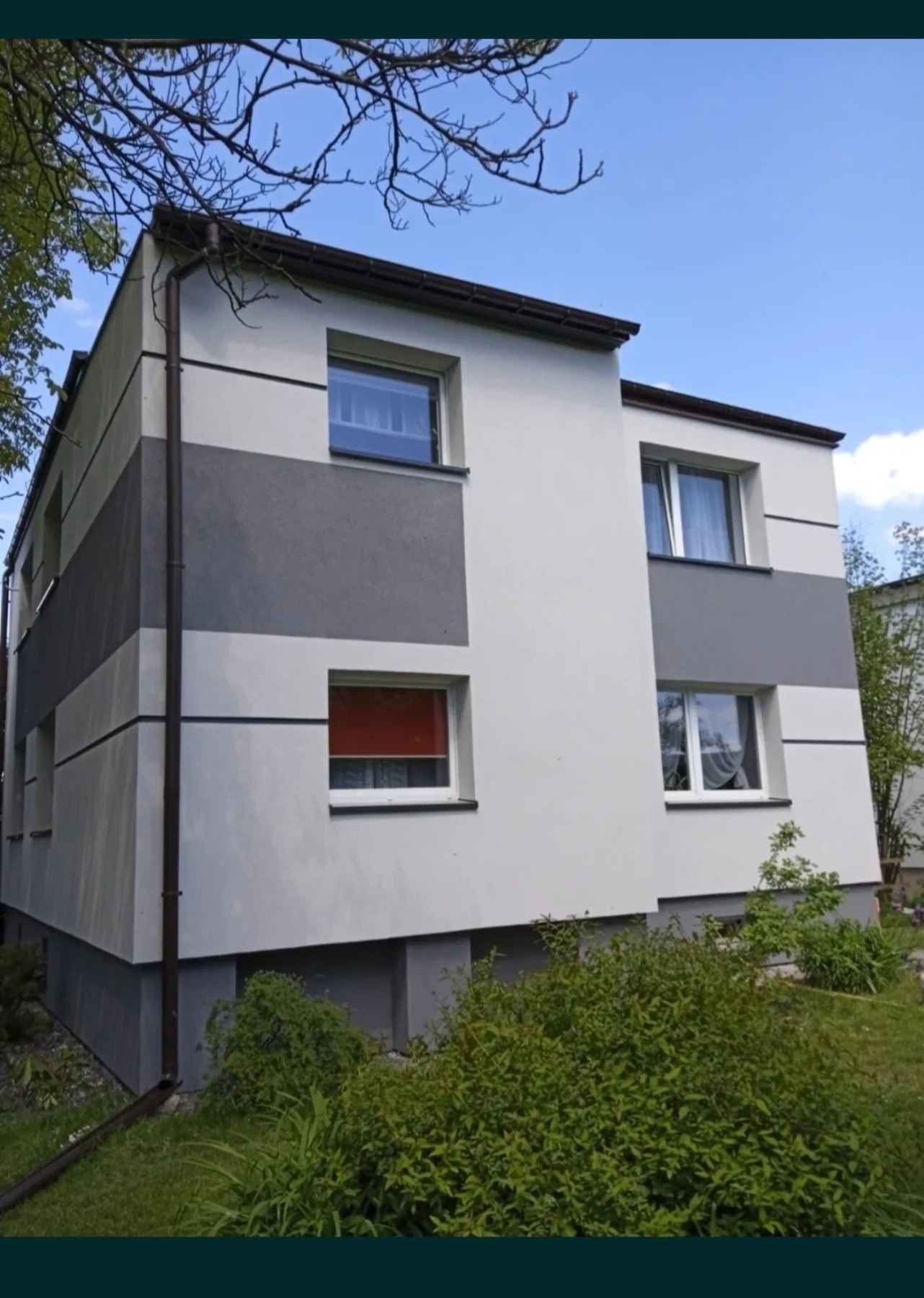 Dwukondygnacyjny budynek z elewacją w odcieniach szarości i bieli, z widocznymi oknami i roślinnością u podstawy. Dom po renowacji z nowym orynnowaniem.