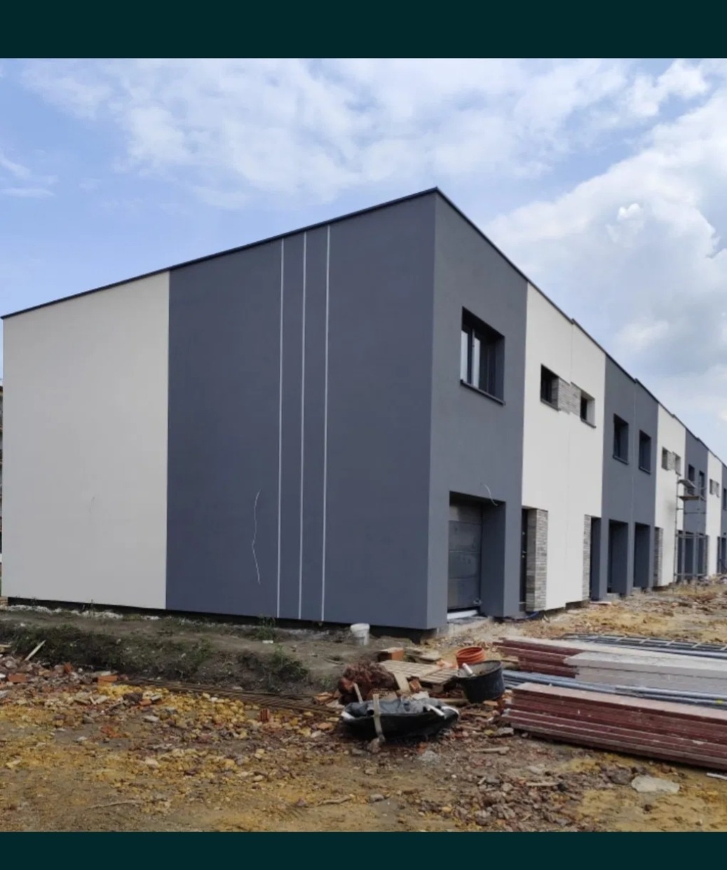 Nowoczesna elewacja szeregowca w odcieniach szarości i bieli z pionowymi zdobieniami. Widoczne materiały budowlane i teren przed budynkiem.