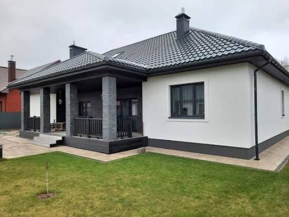 Biały dom z ciemną dachówką i elementami wykończeniowymi, widok z zewnątrz na elewację i fragment ogrodu z młodym drzewkiem.