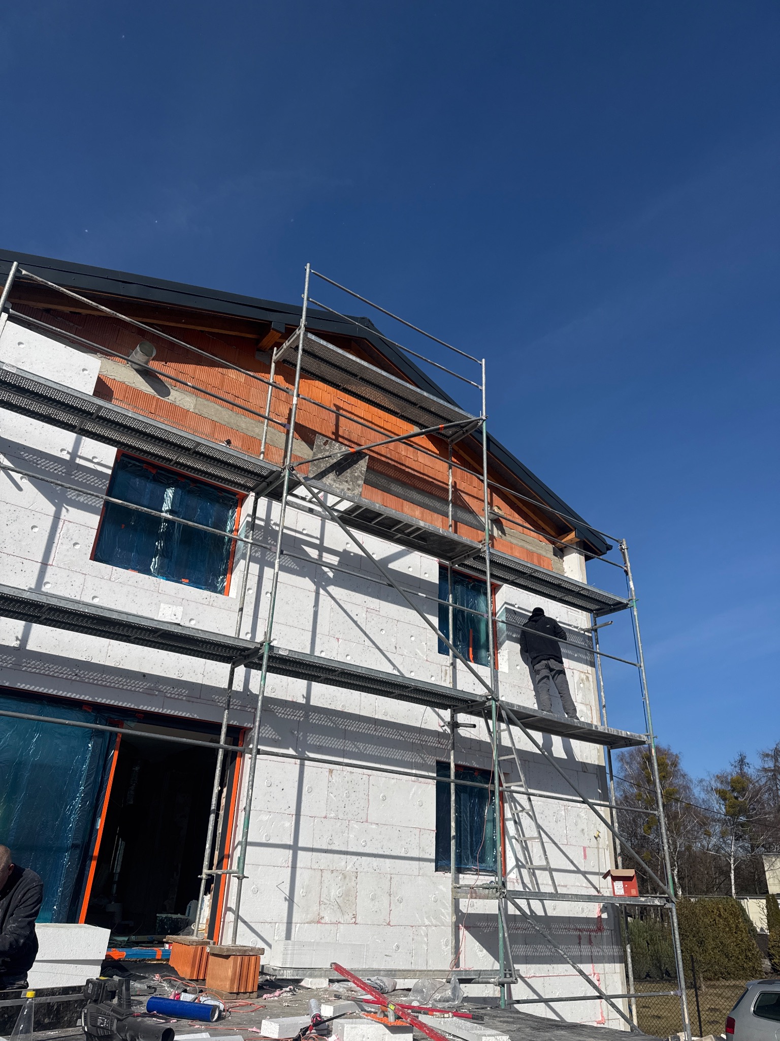 Budowa domu z białych bloczków, widoczne rusztowania i pracownik na nich. Okna zabezpieczone folią. Częściowo wykończona elewacja z cegły klinkierowej.