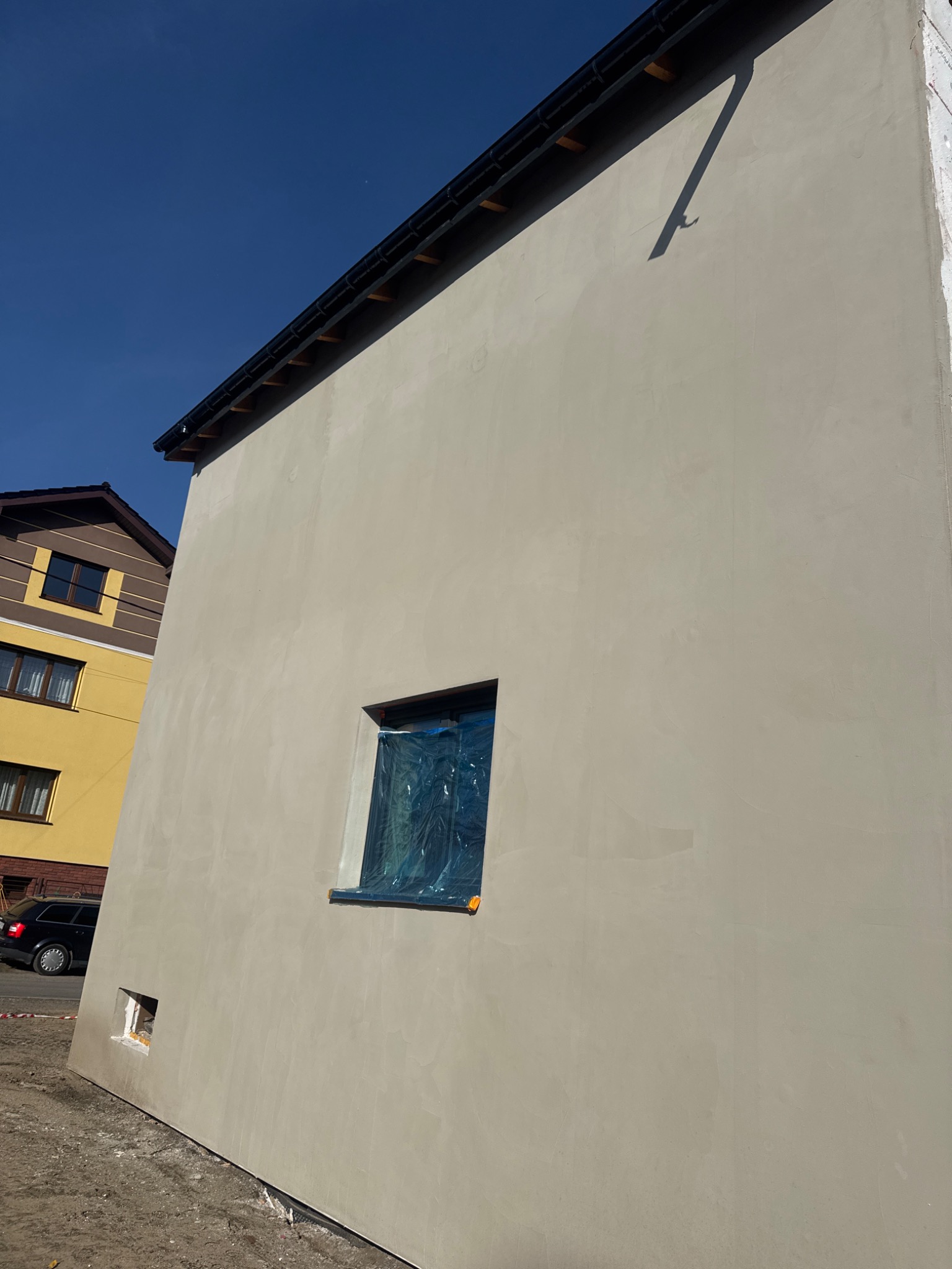 Świeżo otynkowana ściana budynku w neutralnym odcieniu szarości, z zabezpieczonym oknem folią. Widoczny fragment dachu i sąsiedni budynek w tle, błękitne niebo.