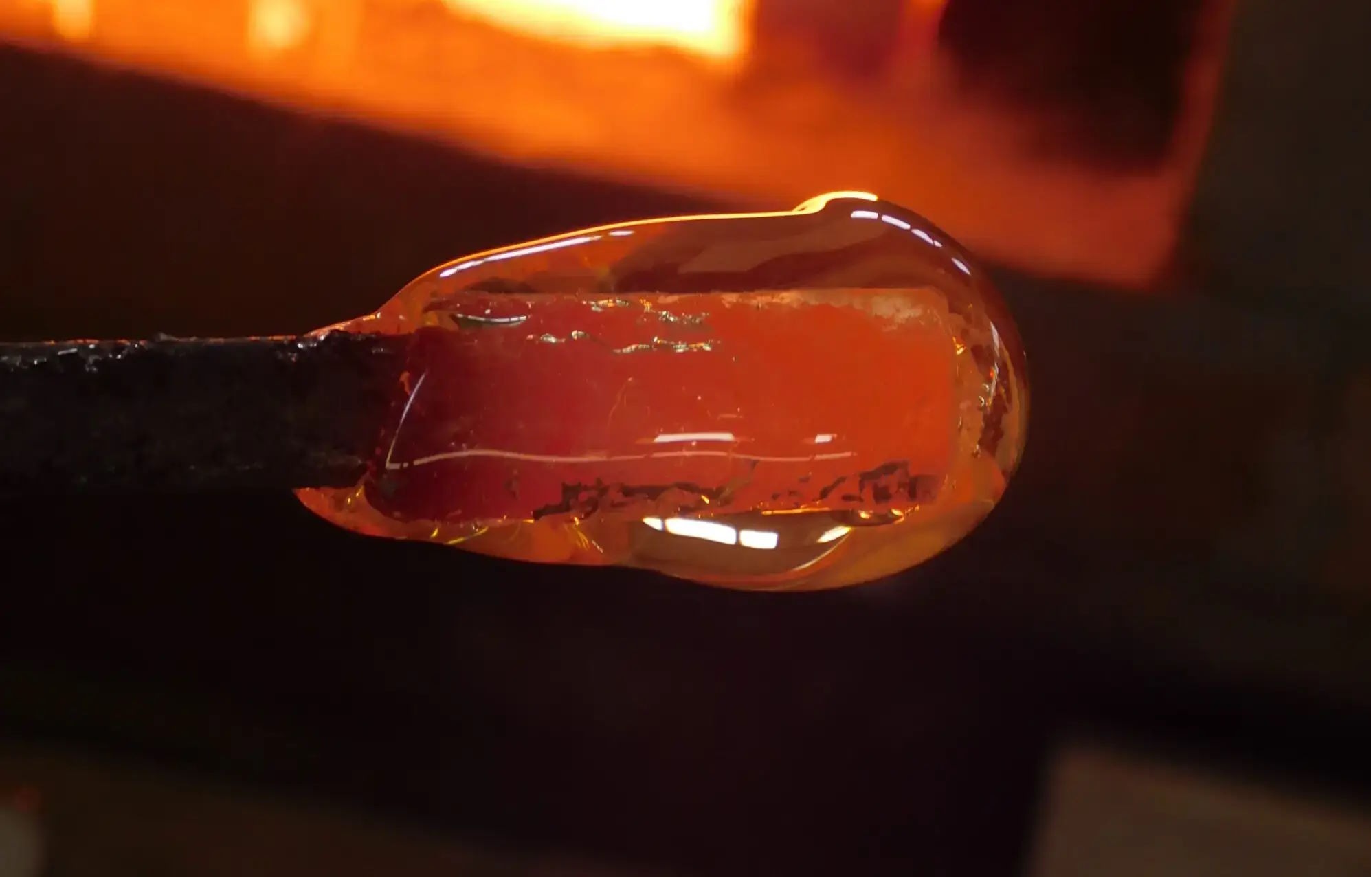 Rozżarzona, formowana masa szklana na czubku narzędzia w warsztacie. Ręczna produkcja szkła, ciepłe światło, ciemne tło. Detal procesu formowania szkła.