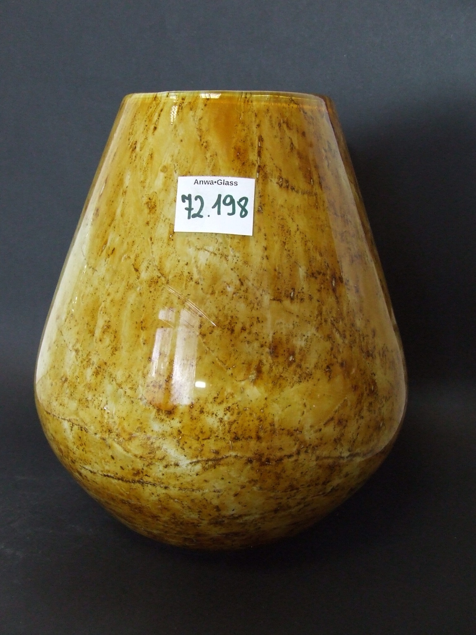 Beżowy, szklany wazon o nieregularnej fakturze, z naklejoną etykietą 'Anwa-Glass 72.198', ustawiony na ciemnym tle.
