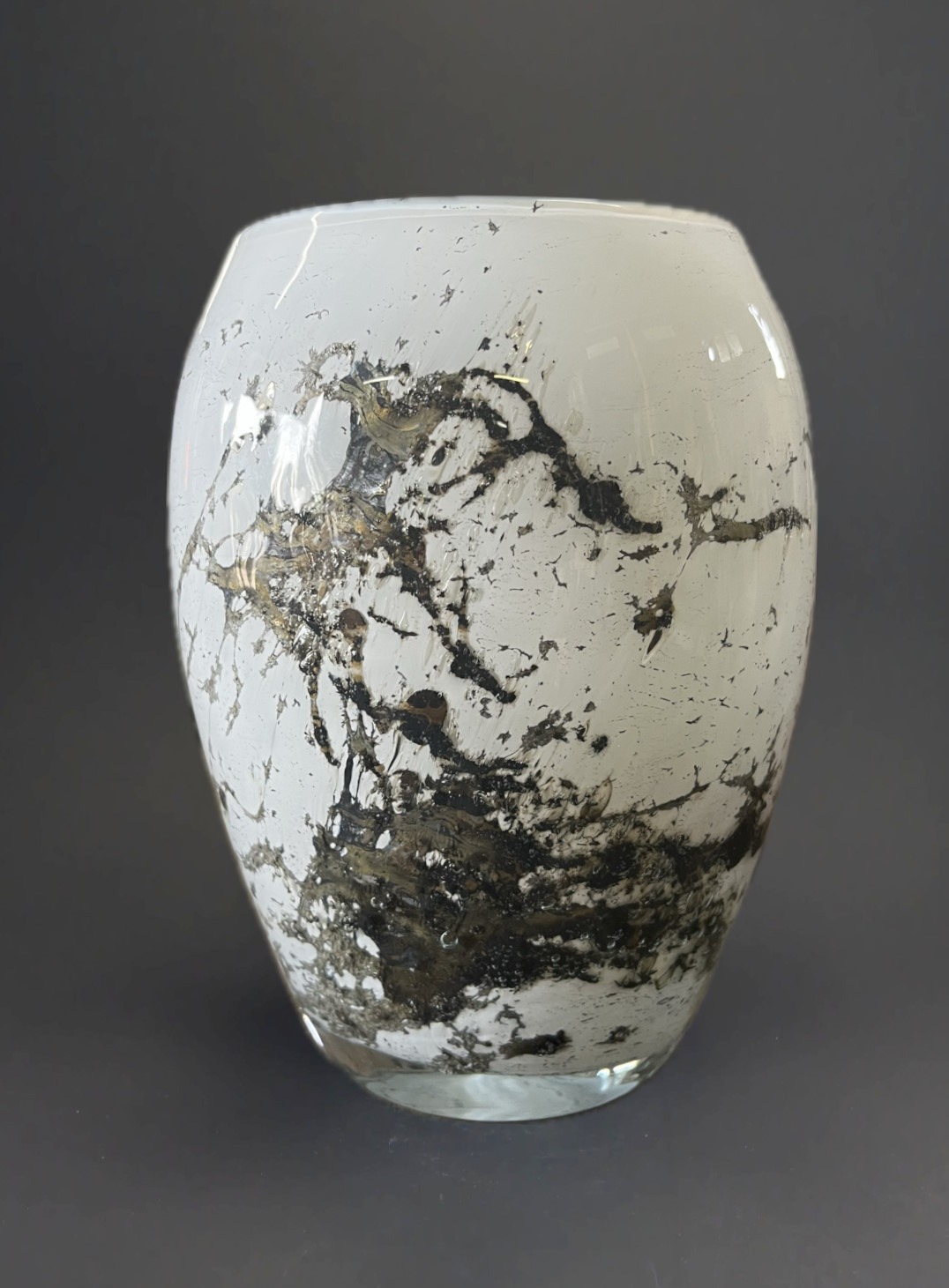 Elegancki, szklany wazon w kolorze białym z czarnymi i złotymi żyłkami, na ciemnoszarym tle. Wazon o obłym kształcie, z widocznymi pęcherzykami powietrza w szkle.