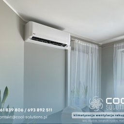 Cool Solutions Dawid Mochocki - Biały klimatyzator ścienny w jasnym pokoju z oknem i firankami, widoczny fragment rośliny doniczkowej. Minimalistyczny design, szare ściany.