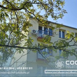Cool Solutions Dawid Mochocki - Klimatyzator na elewacji budynku mieszkalnego, widoczny przez gałęzie drzewa. Logo firmy Cool Solutions w rogu zdjęcia.