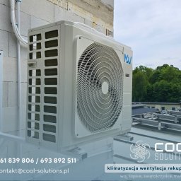 Cool Solutions Dawid Mochocki - Biała jednostka zewnętrzna klimatyzacji zamontowana na ścianie budynku, widoczne przewody i uchwyt montażowy, na tle zieleni i nieba.