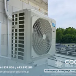 Biała jednostka zewnętrzna klimatyzacji zamontowana na ścianie budynku, widoczne przewody i uchwyt montażowy, na tle zieleni i nieba.