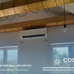 Biała klimatyzacja ścienna w jasnym wnętrzu z drewnianymi belkami stropowymi i wiszącą żarówką. Widoczne okna i elementy dekoracyjne.