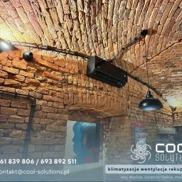 Cool Solutions Dawid Mochocki - Czarny klimatyzator zamontowany na ceglanym, łukowym suficie w lokalu, z widocznymi przewodami instalacji i oświetleniem. W tle bar z wyposażeniem.