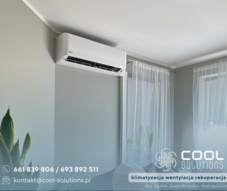 Biały klimatyzator ścienny w jasnym pokoju z oknem i firankami, widoczny fragment rośliny doniczkowej. Minimalistyczny design, szare ściany.