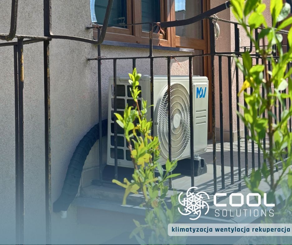 Zamontowana jednostka zewnętrzna klimatyzacji na balkonie w Goleszowie. Widoczny czarny peszel i fragment balustrady. Częściowo zasłonięte przez roślinę.