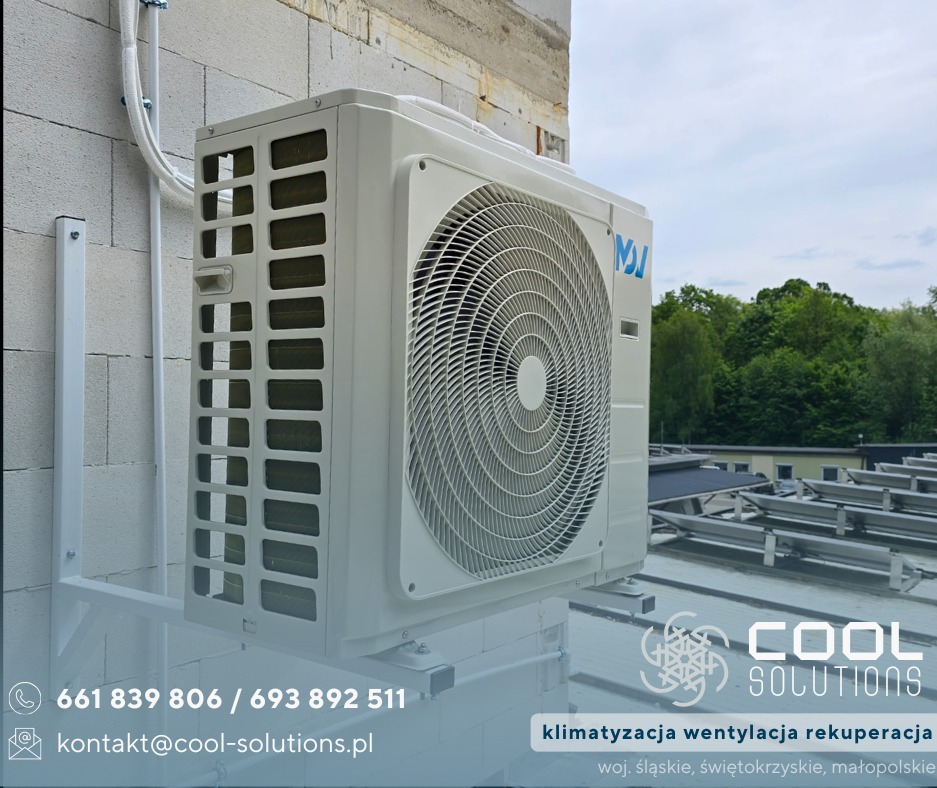 Biała jednostka zewnętrzna klimatyzacji zamontowana na ścianie budynku, widoczne przewody i uchwyt montażowy, na tle zieleni i nieba.