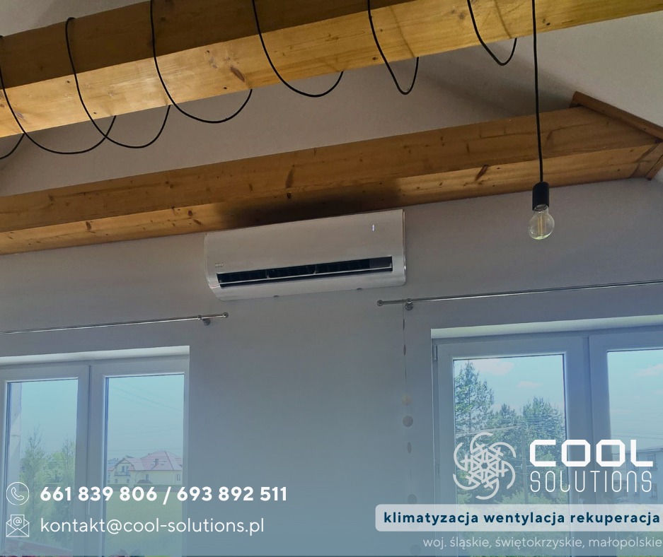 Biała klimatyzacja ścienna w jasnym wnętrzu z drewnianymi belkami stropowymi i wiszącą żarówką. Widoczne okna i elementy dekoracyjne.