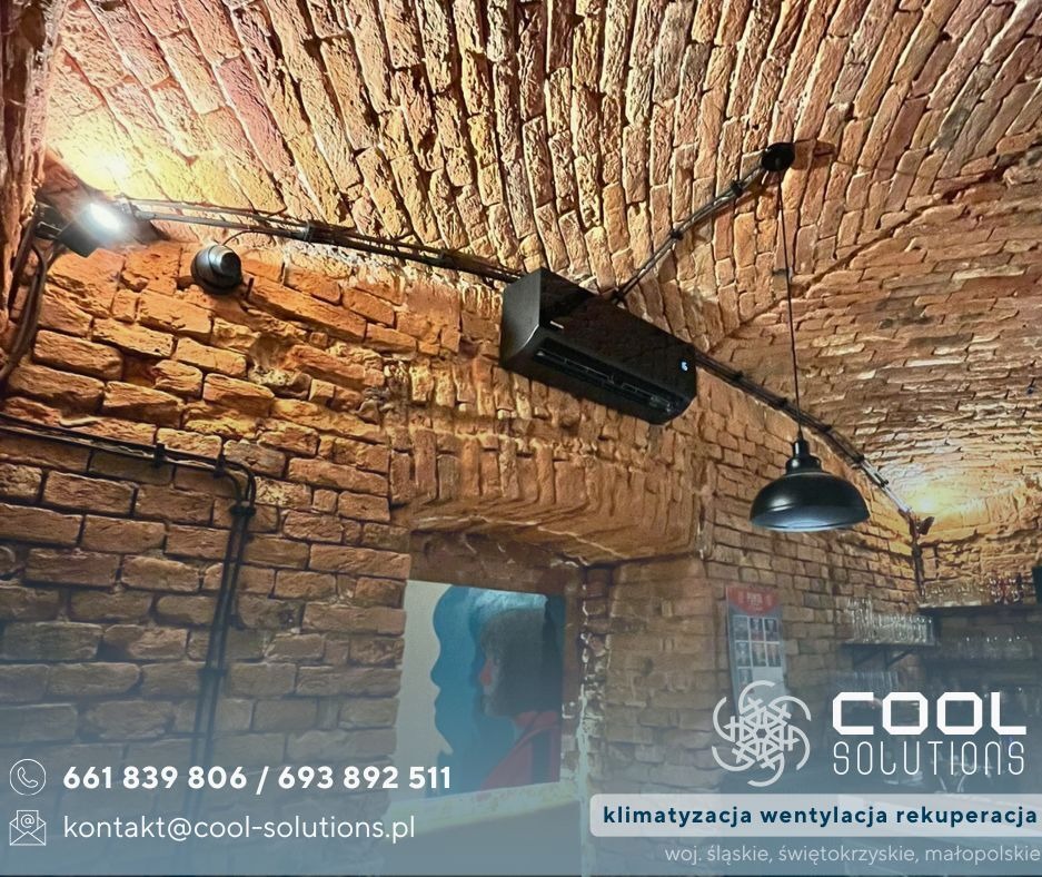 Czarny klimatyzator zamontowany na ceglanym, łukowym suficie w lokalu, z widocznymi przewodami instalacji i oświetleniem. W tle bar z wyposażeniem.