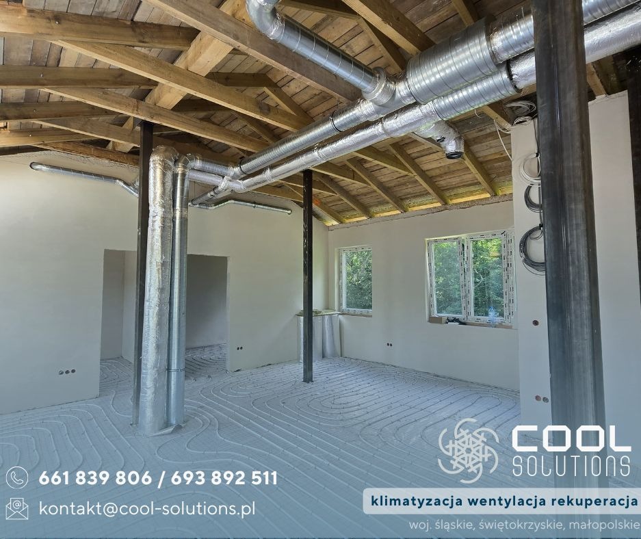 Wnętrze w trakcie montażu instalacji wentylacyjnej z widocznymi rurami pod sufitem i ogrzewaniem podłogowym. Surowy stan wykończenia, drewniana konstrukcja dachu.
