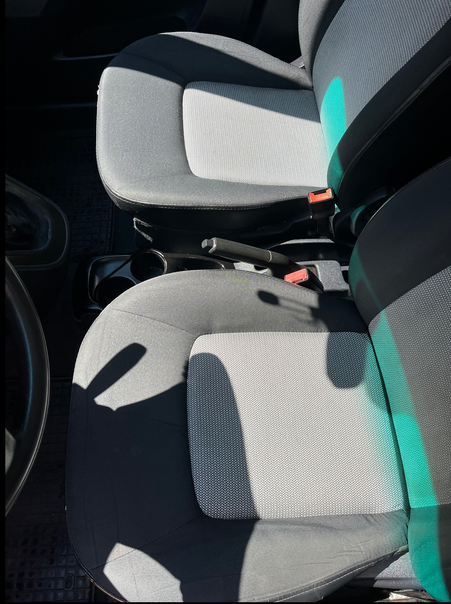 Szare, tekstylne fotele samochodowe z widocznymi pasami bezpieczeństwa i elementami deski rozdzielczej, oświetlone naturalnym światłem słonecznym.