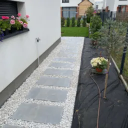 Ścieżka z płyt betonowych i białego grysu w nowoczesnym ogrodzie. Donica z kwiatami na parapecie, krzewy przy ogrodzeniu. Czarna agrowłóknina pod roślinami.