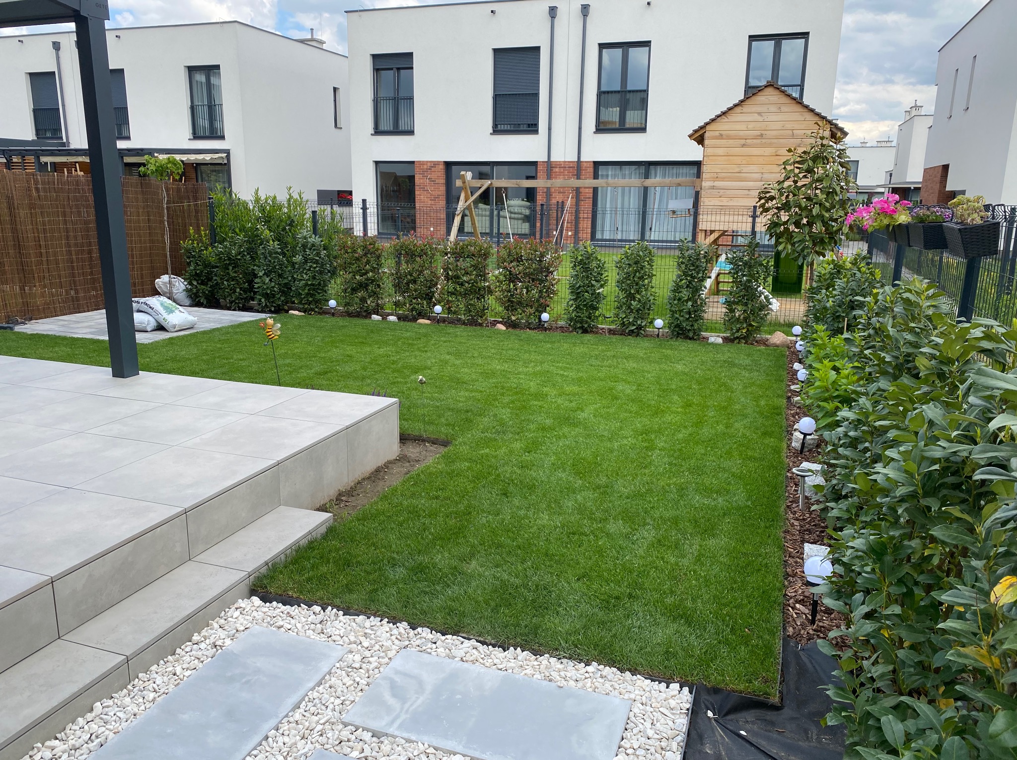 Zadbany ogród z równo przystrzyżonym trawnikiem, ozdobnymi kamieniami i płytami chodnikowymi. W tle nowoczesny dom z placem zabaw i drewnianym domkiem.