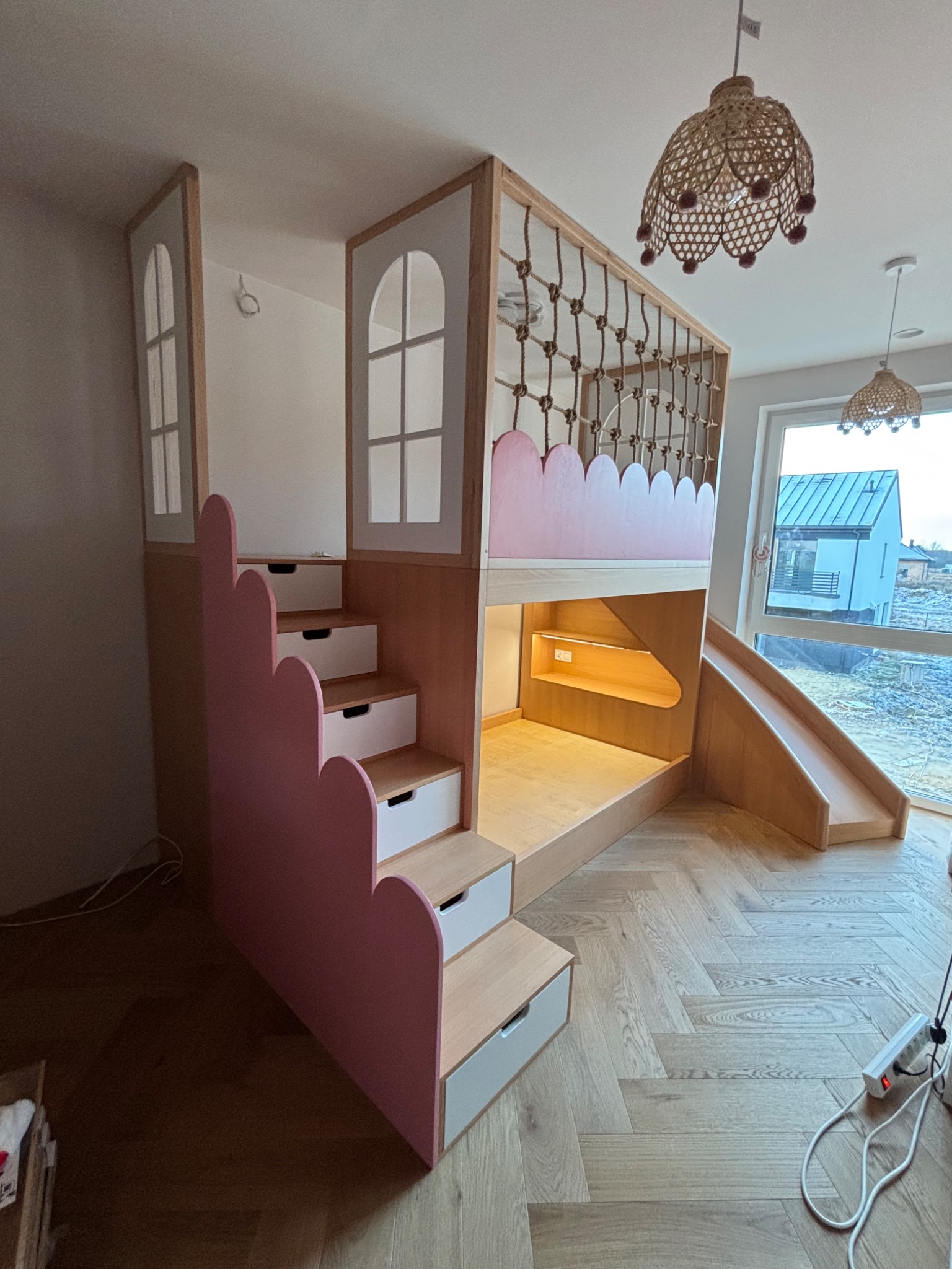 Drewniane łóżko piętrowe dla dziecka z schodami w kształcie chmurki i zjeżdżalnią, oświetlone lampami z wikliny, na podłodze parkiet w jodełkę.