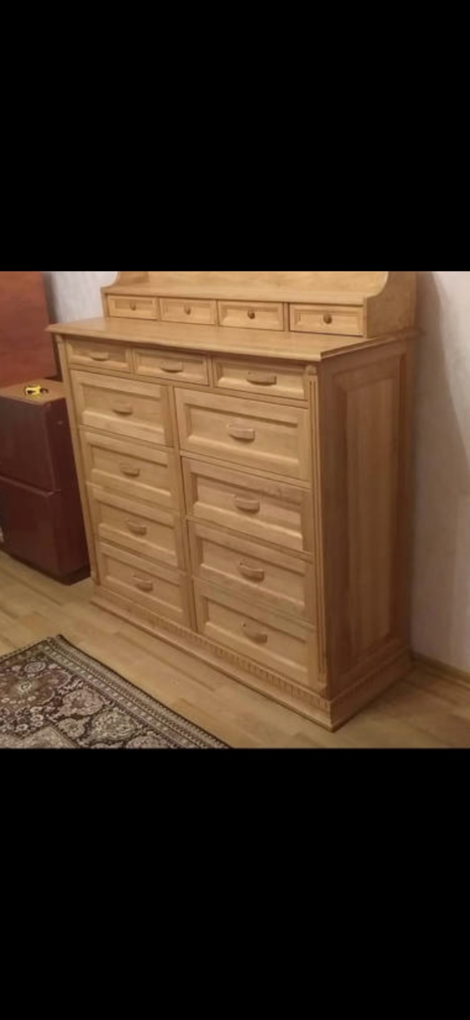 Jasny, drewniany komoda z szufladami, widoczna na tle dywanu i ściany. Mebel o klasycznym designie, z delikatnymi zdobieniami i uchwytami.