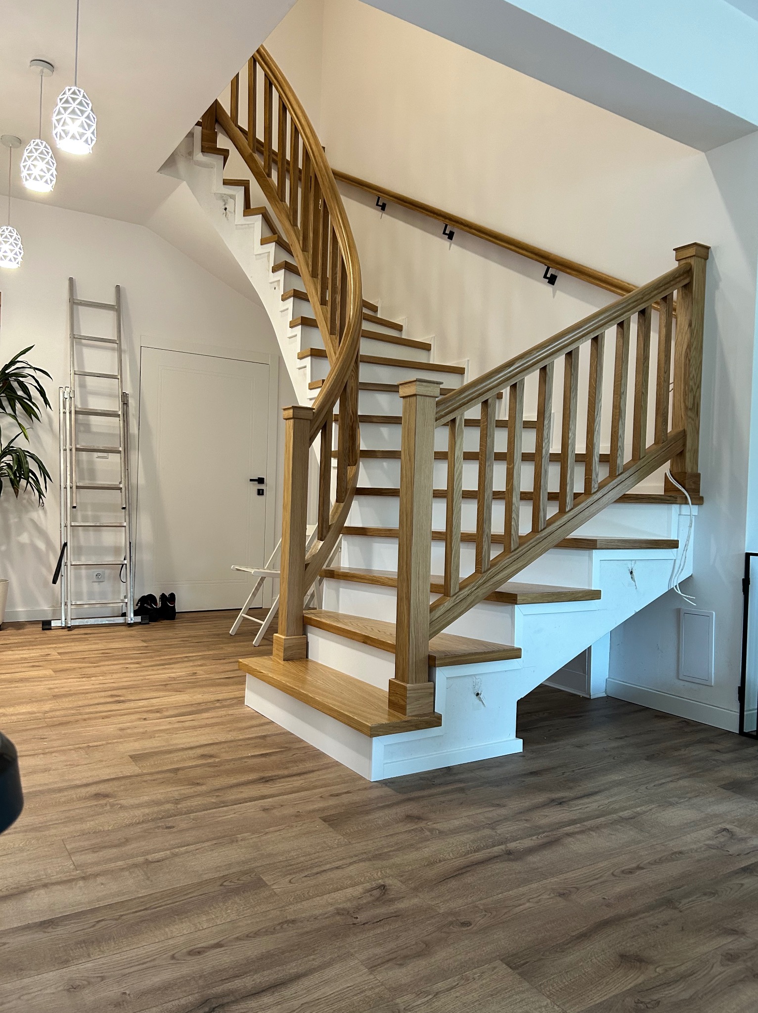 Jasne, drewniane schody z białymi podstopnicami w nowoczesnym wnętrzu. Widoczne lampy wiszące, drabina i drewniana podłoga. Ujęcie z dołu, podkreślające zakrzywiony kształt.