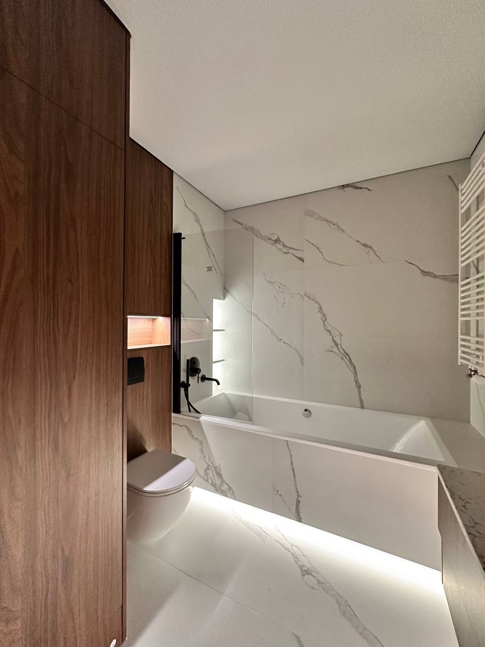 Nowoczesna łazienka z marmurowymi płytkami, podświetlaną wanną i drewnianą zabudową. Minimalistyczny design, czarne akcenty armatury i oświetlenie LED.