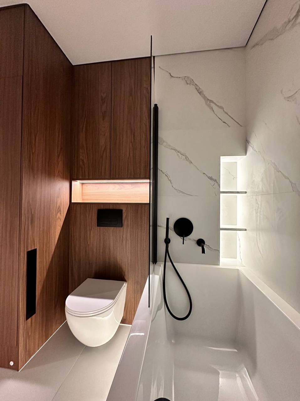 Nowoczesna łazienka z drewnianą zabudową, białą wanną i toaletą. Marmurowe płytki z podświetlanymi półkami. Czarna armatura i minimalistyczny design.