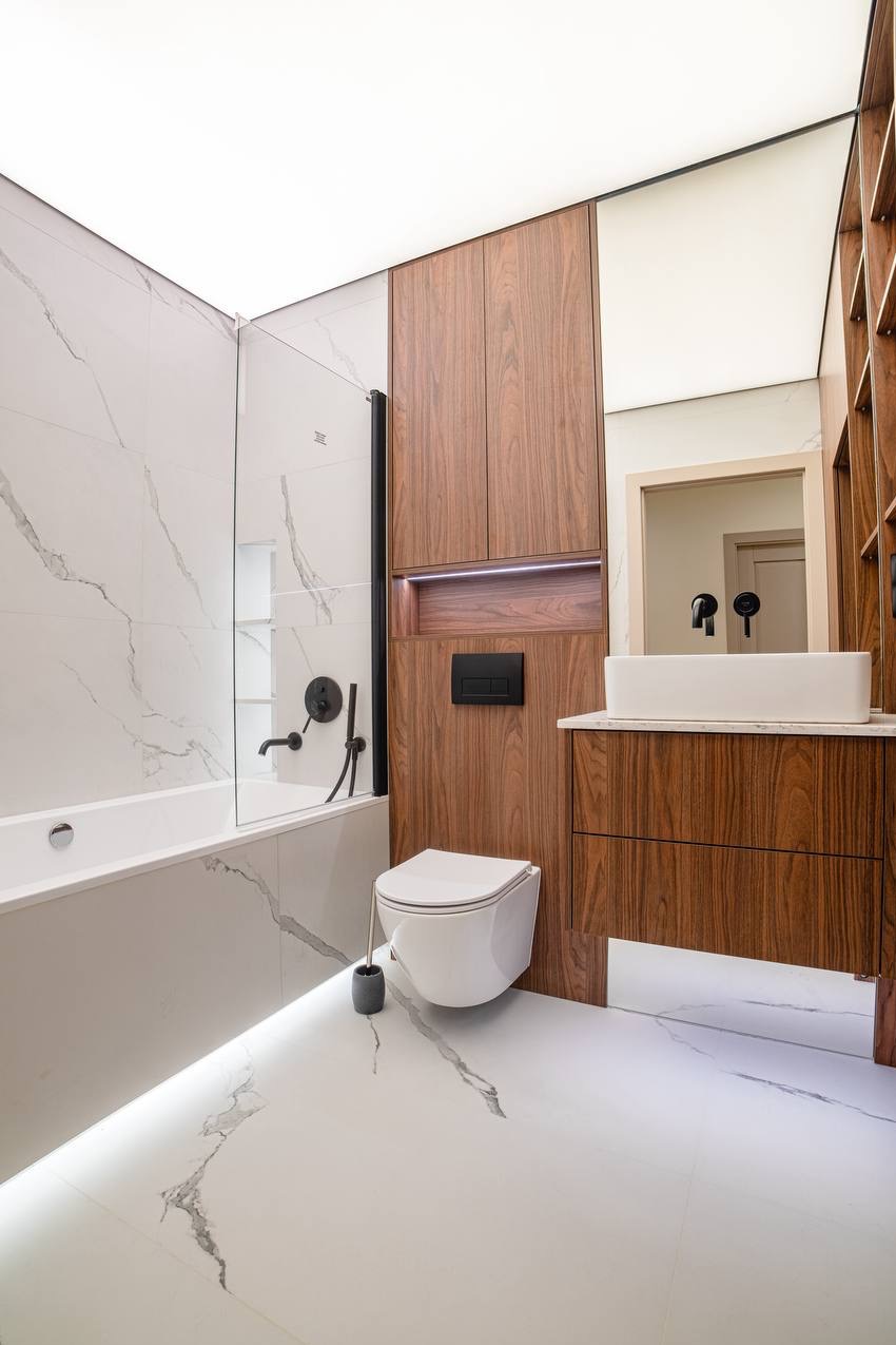 Nowoczesna łazienka z białymi płytkami imitującymi marmur, drewnianymi szafkami i czarną armaturą. Podświetlenie LED pod wanną i szafką dodaje elegancji.