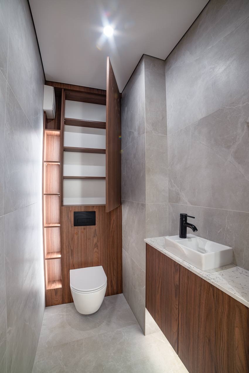 Nowoczesna łazienka z szarymi płytkami, drewnianą zabudową WC i szafką pod umywalką z blatem imitującym marmur. Minimalistyczna armatura i oświetlenie.