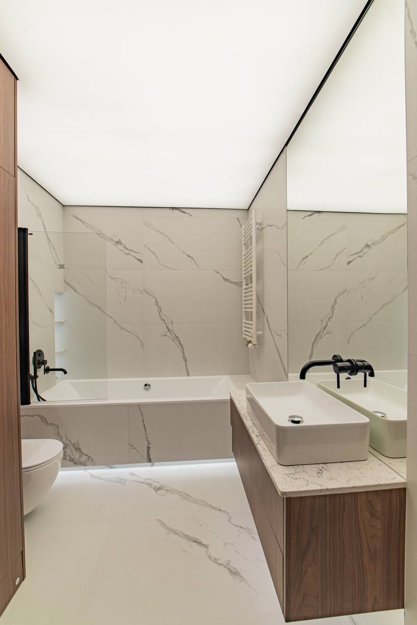 Nowoczesna łazienka z białymi płytkami imitującymi marmur, podświetlana wanna i umywalka nablatowa z czarną armaturą. Minimalistyczny design z drewnianymi akcentami.