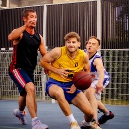 Spizoczko - Trzech koszykarzy w akcji podczas meczu na zewnątrz. Jeden w żółtej koszulce z piłką, drugi w czarnej, trzeci w białej. W tle budynek i ogrodzenie.