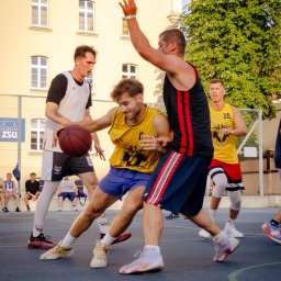 Spizoczko - Mecz koszykówki ulicznej: dynamiczna akcja zawodników w sportowych strojach na tle żółtego budynku z oknami i zieleni drzew. Ujęcie z bliska, w ruchu.