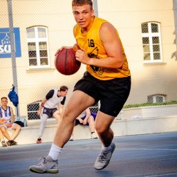 Spizoczko - Młody koszykarz w żółtej koszulce nr 16 dynamicznie prowadzi piłkę na niebieskim boisku, w tle budynek z oknami i siatka. Gra w koszykówkę w słoneczny dzień.