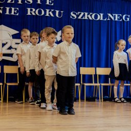 Spizoczko - Uczniowie ustawieni w rzędzie na tle dekoracji z okazji zakończenia roku szkolnego 2025. Chłopcy w białych koszulach, dziewczynki w białych bluzkach i granatowych spódnicach.