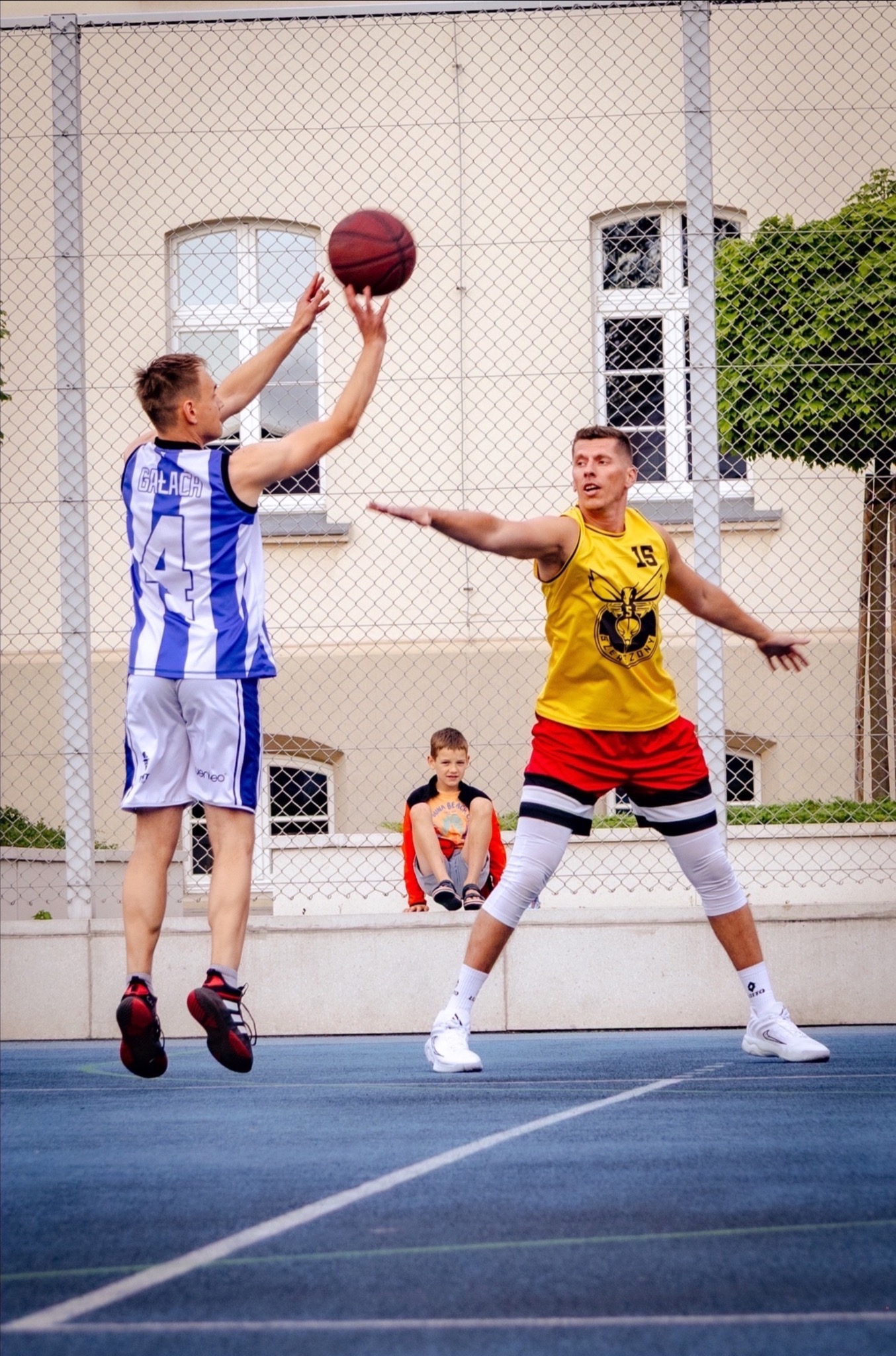 Dynamiczne ujęcie: koszykarz w locie oddaje rzut, drugi zawodnik w gotowości do obrony, a w tle chłopiec obserwuje grę na tle ogrodzenia i budynku.