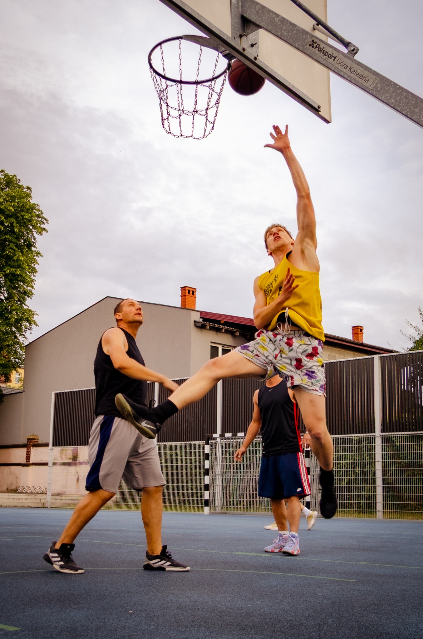 Mężczyzna w żółtej koszulce skacze do kosza podczas gry w koszykówkę na zewnątrz. Widoczni inni gracze i budynki w tle.