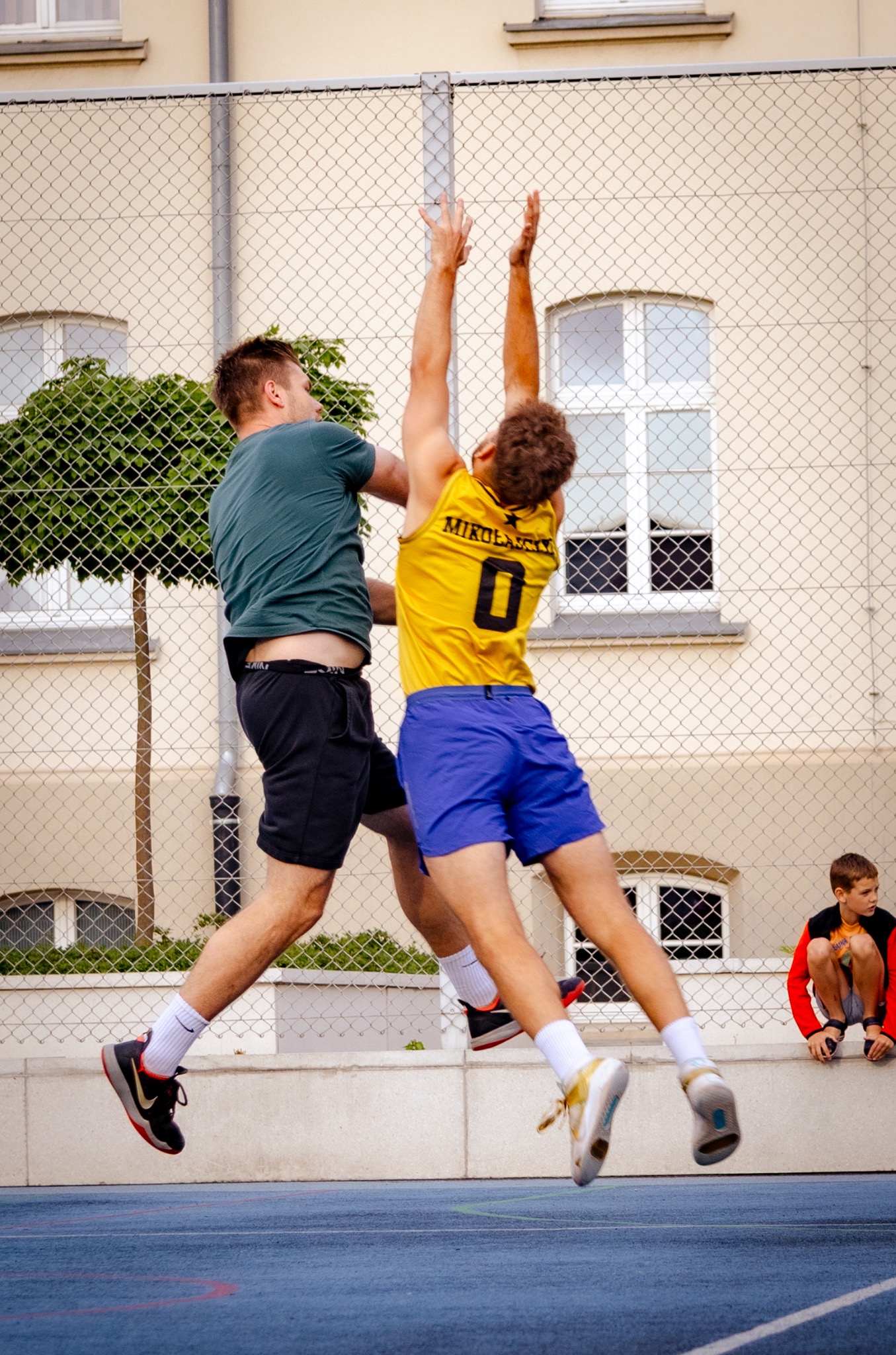 Dwóch młodych mężczyzn skacze, rywalizując o piłkę na tle budynku z siatką. Jeden ubrany w żółtą koszulkę z numerem '0', drugi w zieloną. Widoczny obserwator.