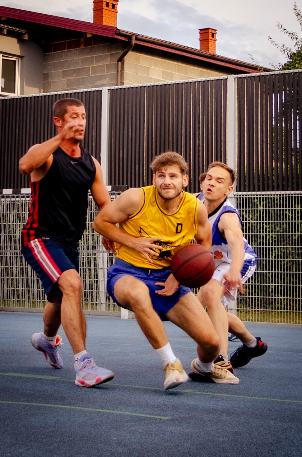 Trzech koszykarzy w akcji podczas meczu na zewnątrz. Jeden w żółtej koszulce z piłką, drugi w czarnej, trzeci w białej. W tle budynek i ogrodzenie.