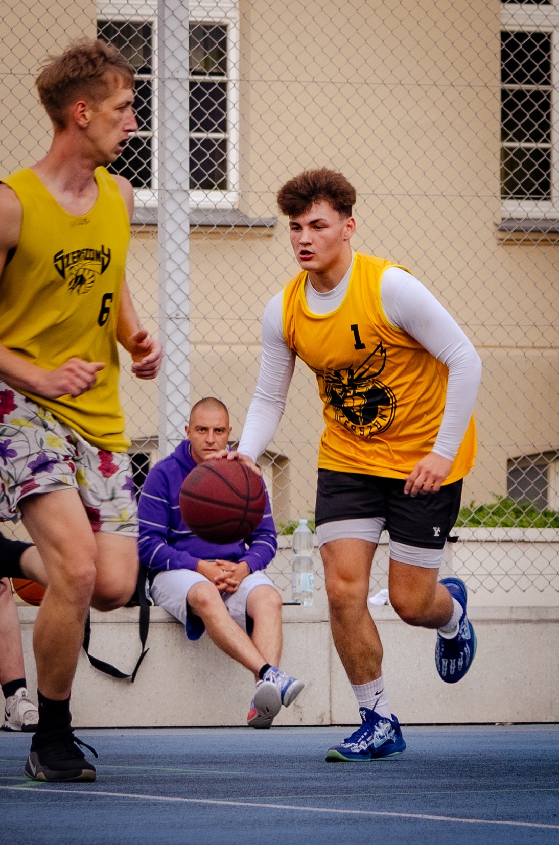 Młody koszykarz w żółtej koszulce z numerem 1 w akcji na boisku; w tle zawodnik z numerem 6 i siedzący mężczyzna z piłką. Scena z meczu amatorskiego.