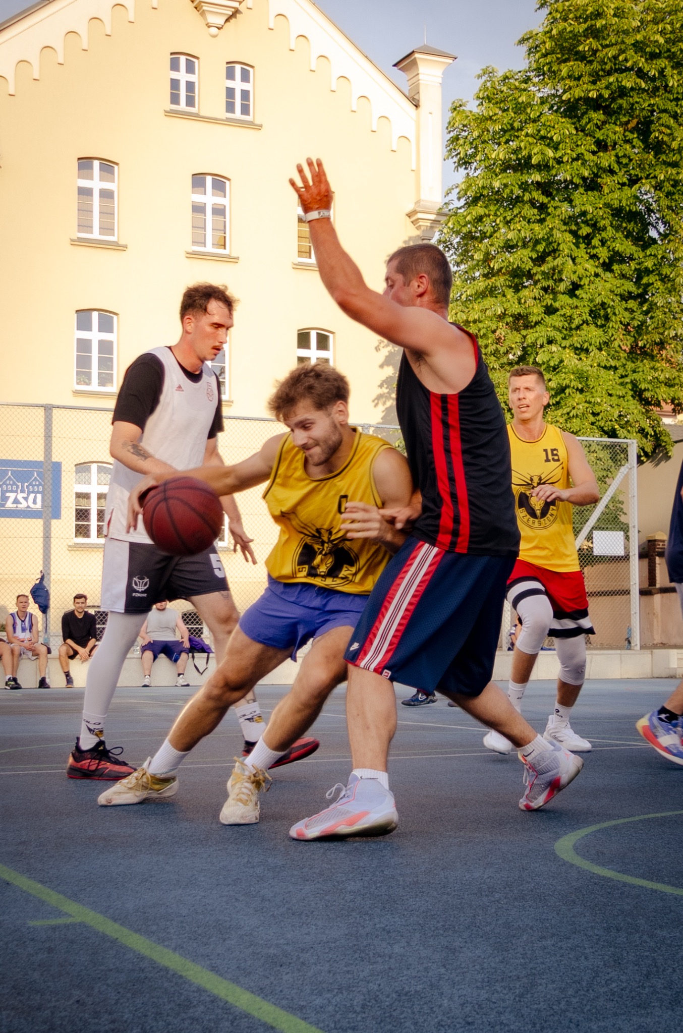 Mecz koszykówki ulicznej: dynamiczna akcja zawodników w sportowych strojach na tle żółtego budynku z oknami i zieleni drzew. Ujęcie z bliska, w ruchu.