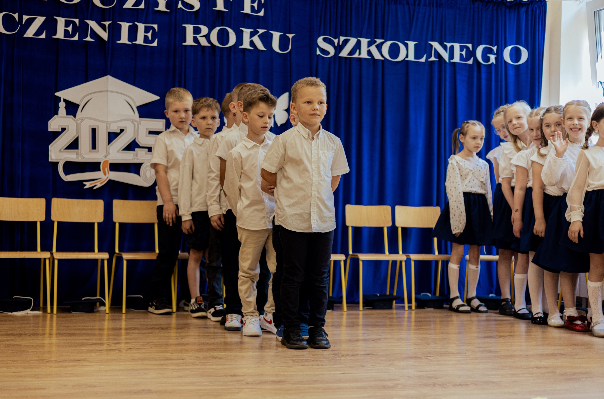 Uczniowie ustawieni w rzędzie na tle dekoracji z okazji zakończenia roku szkolnego 2025. Chłopcy w białych koszulach, dziewczynki w białych bluzkach i granatowych spódnicach.