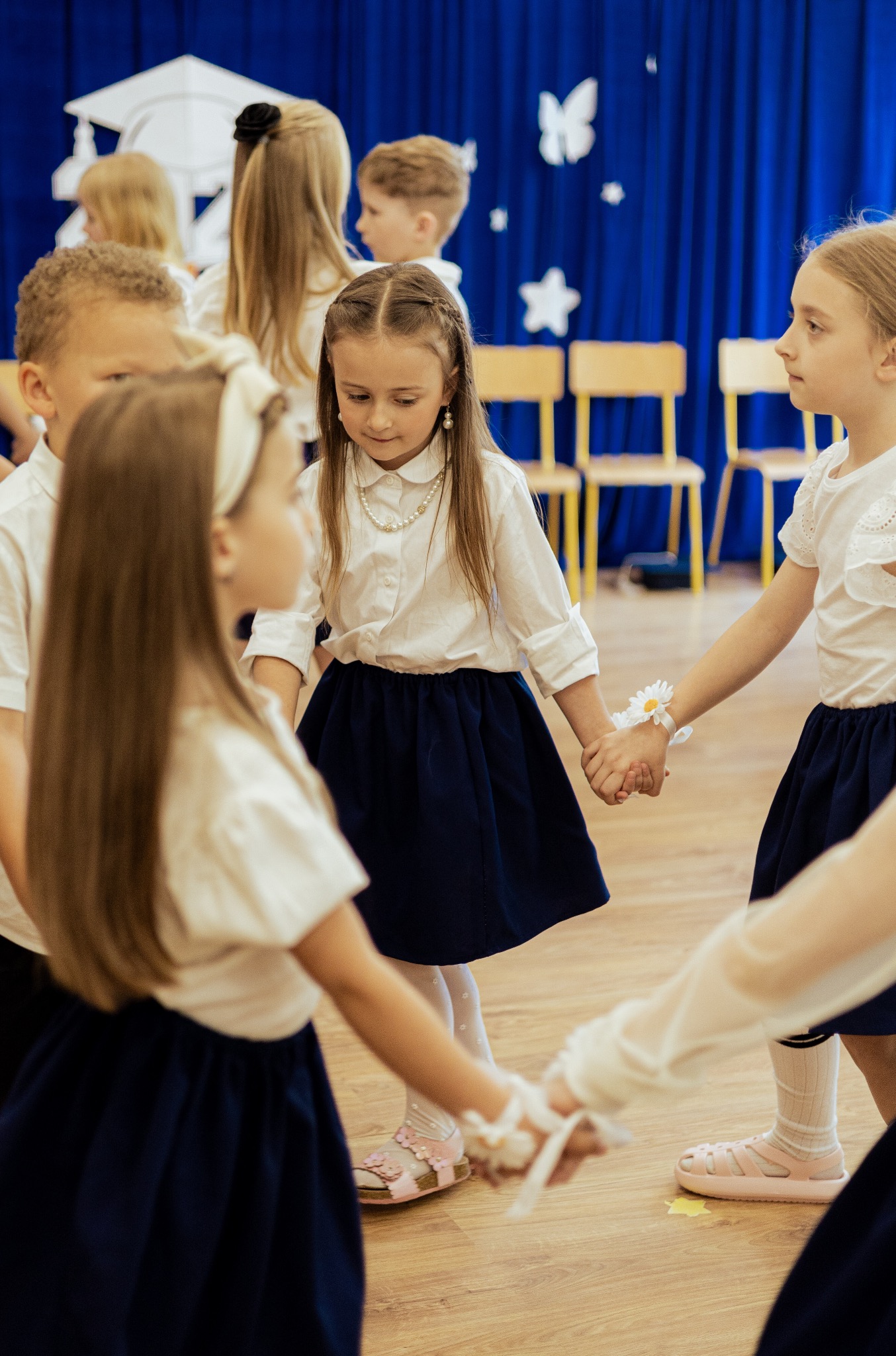 Dzieci w białych koszulach i granatowych spódniczkach trzymają się za ręce, tworząc krąg na tle niebieskiej dekoracji z motywami gwiazd i motyli.