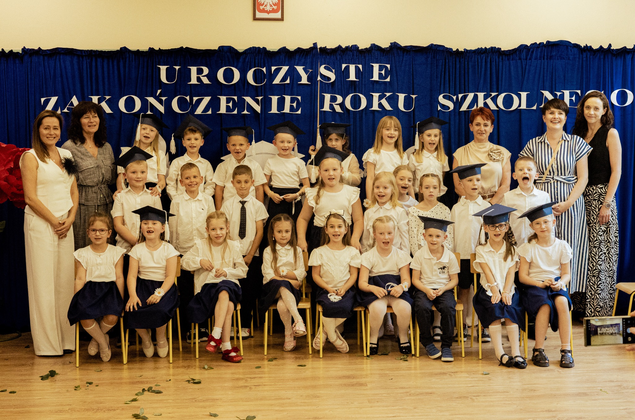 Grupowe zdjęcie uczniów w togach i biretach podczas uroczystości zakończenia roku szkolnego, z nauczycielami na tle niebieskiej kotary. Radosne uśmiechy i konfetti na podłodze.