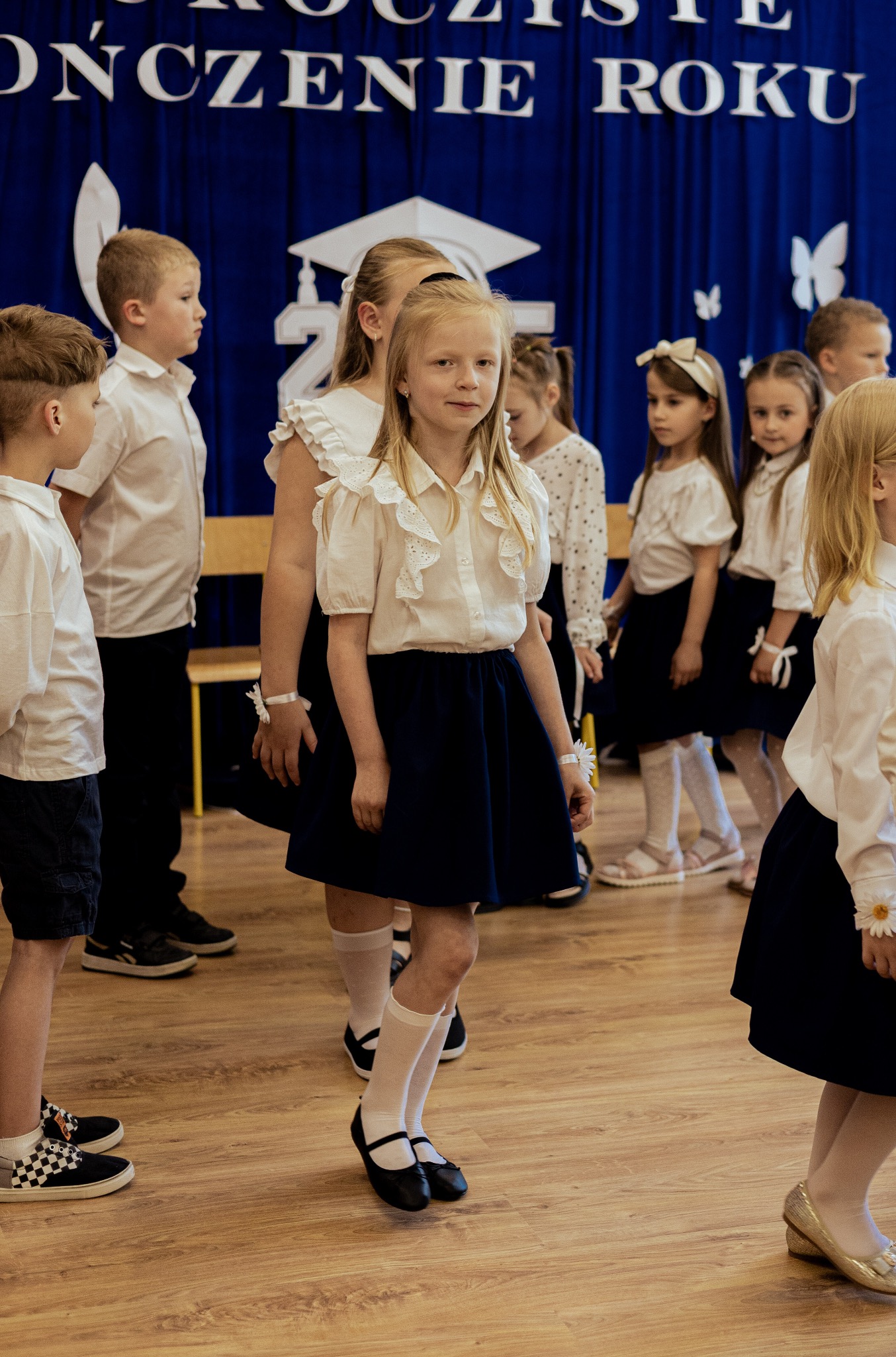 Uczniowie w białych bluzkach i granatowych spódnicach/spodenkach stoją na drewnianej podłodze na tle dekoracji z okazji zakończenia roku szkolnego.
